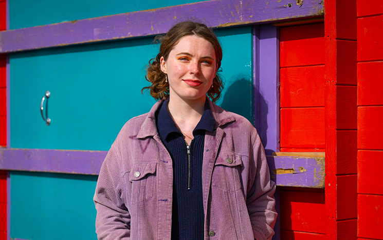 Student smiling at camera standing in front of colourful beach hut